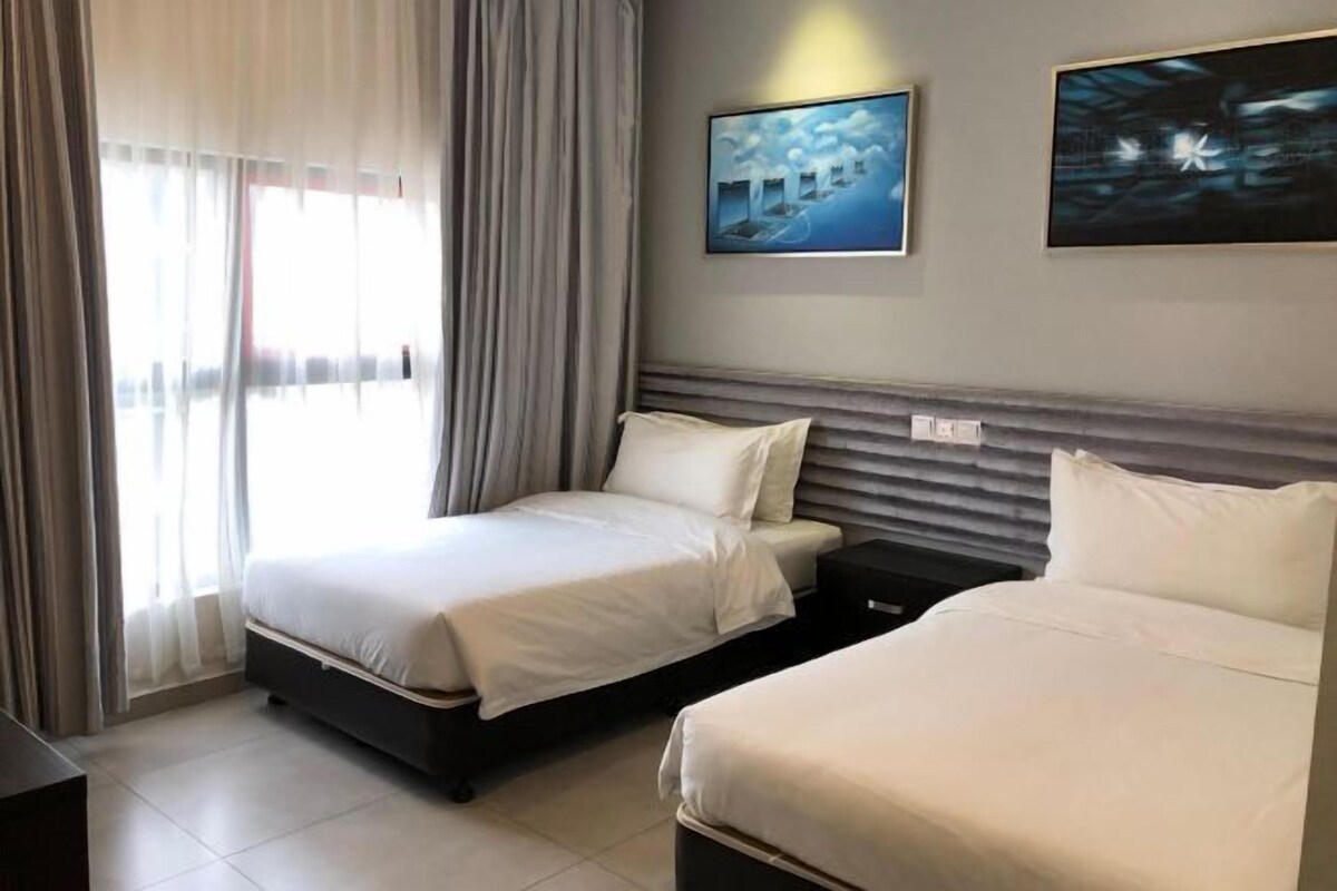 A hotel room with two beds, a large window with curtains, and two framed artworks on the wall.