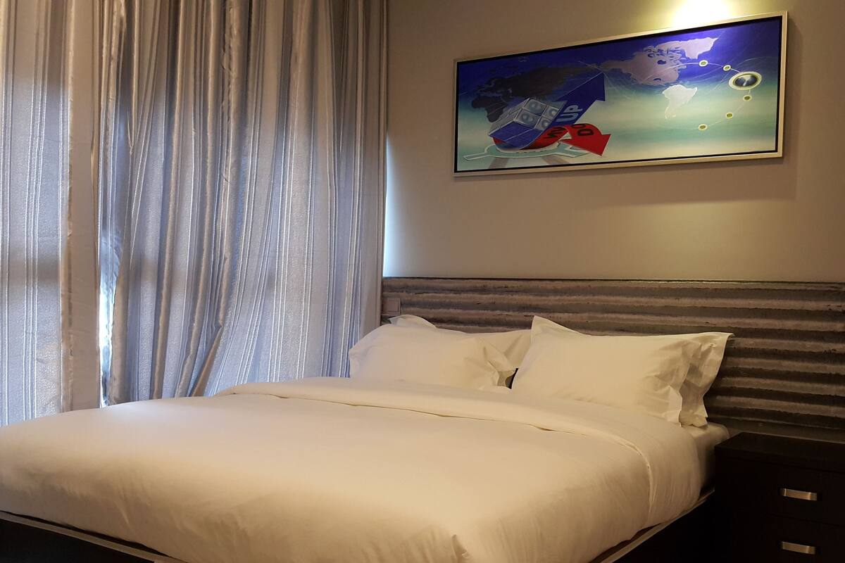 A hotel room with a neatly made bed, a headboard with horizontal stripes, and a painting of a globe and dice on the wall.