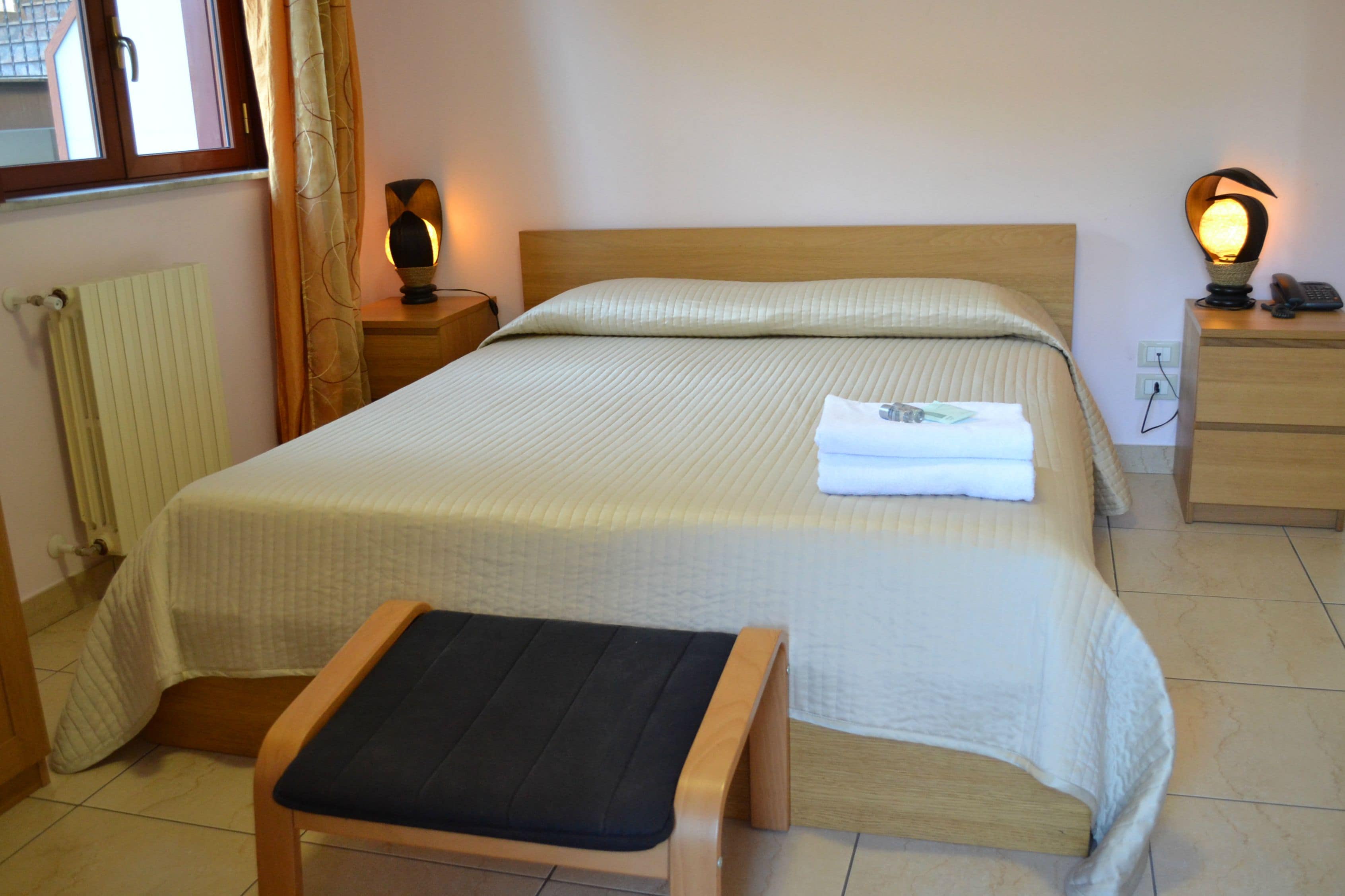 Una cama bien hecha con cabecera de madera, una mesita de noche con lámpara, y un banco al pie de la cama.