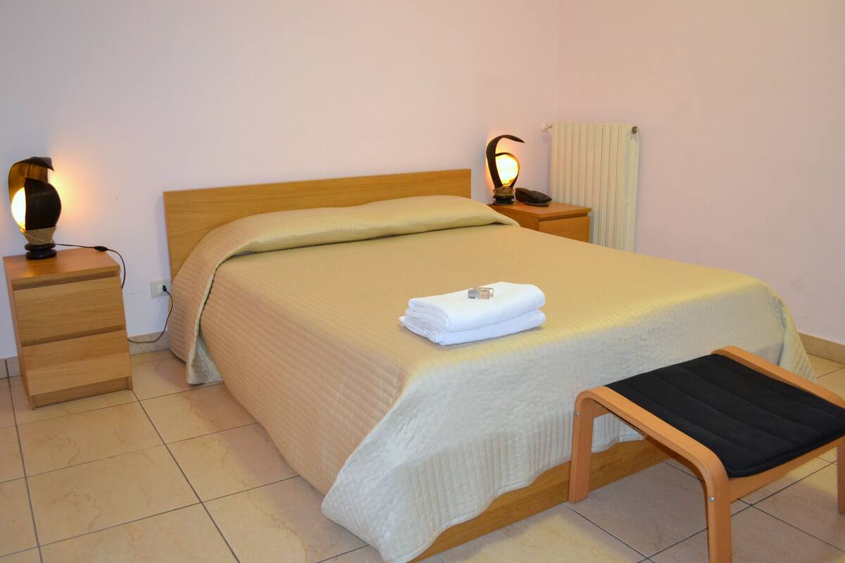 A neatly made bed with a folded towel, a bedside table with a lamp, and a small wooden stool.