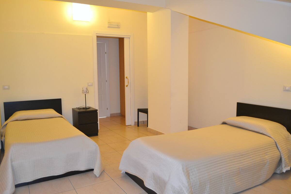 A hotel room with two beds, a bedside table, a chair, and a door leading to another room.