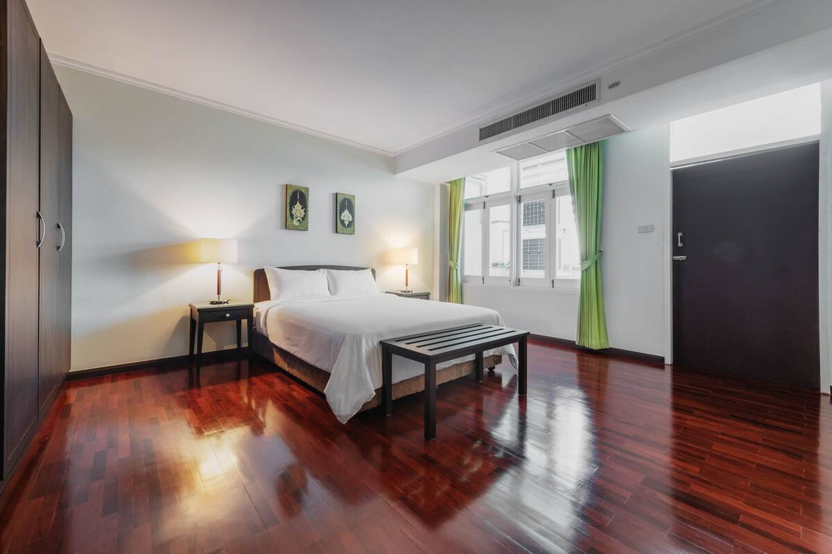 A hotel room with a wooden floor, a bed with white bedding, two bedside tables with lamps, and a window with green curtains.