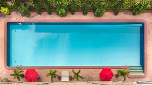 A rectangular pool with a clear blue water surface, surrounded by a pink wall and a row of green plants. There are two red umbrellas and a few small palm trees at the pool's edge. - Capital Mansion (Bangkok)