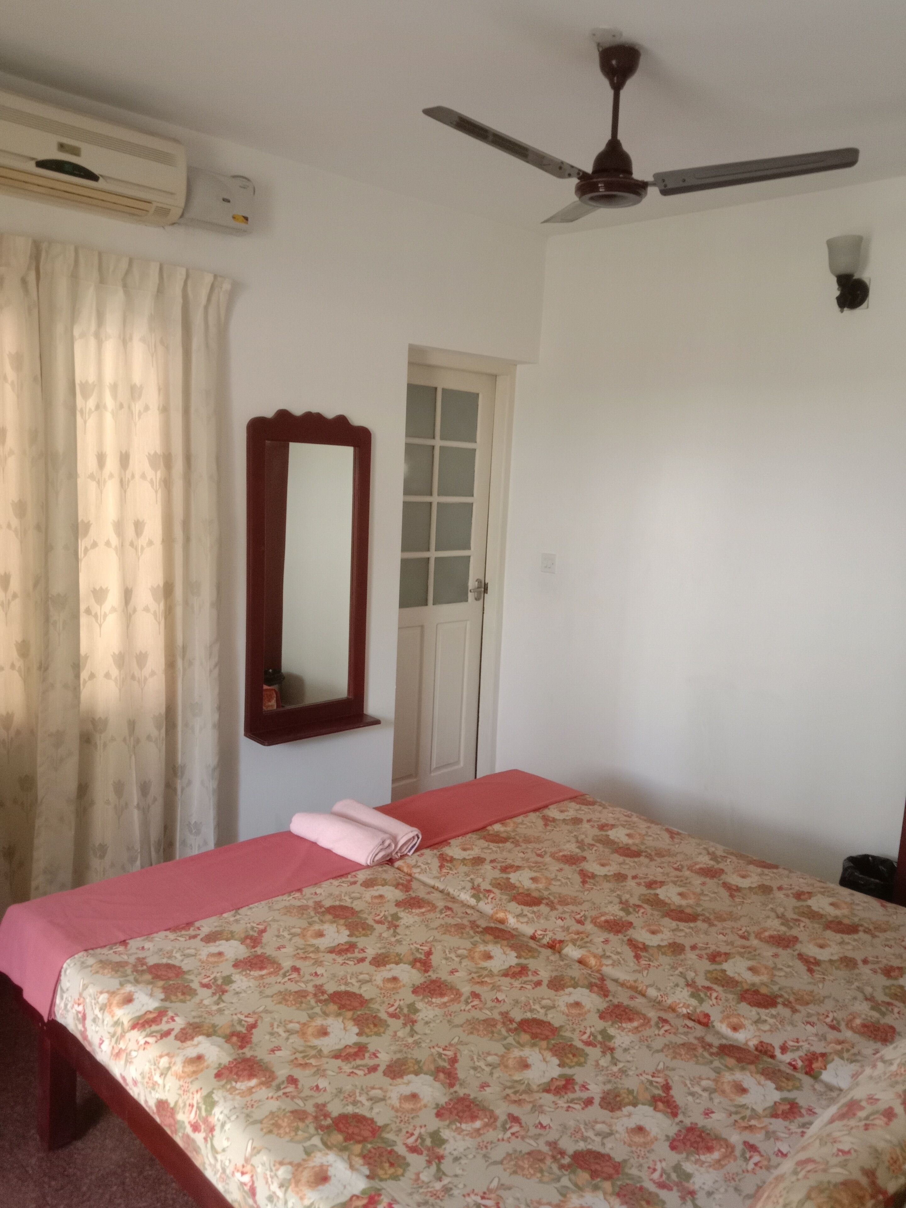 A bedroom with a bed, a ceiling fan, an air conditioning unit, a door, and a window with curtains.