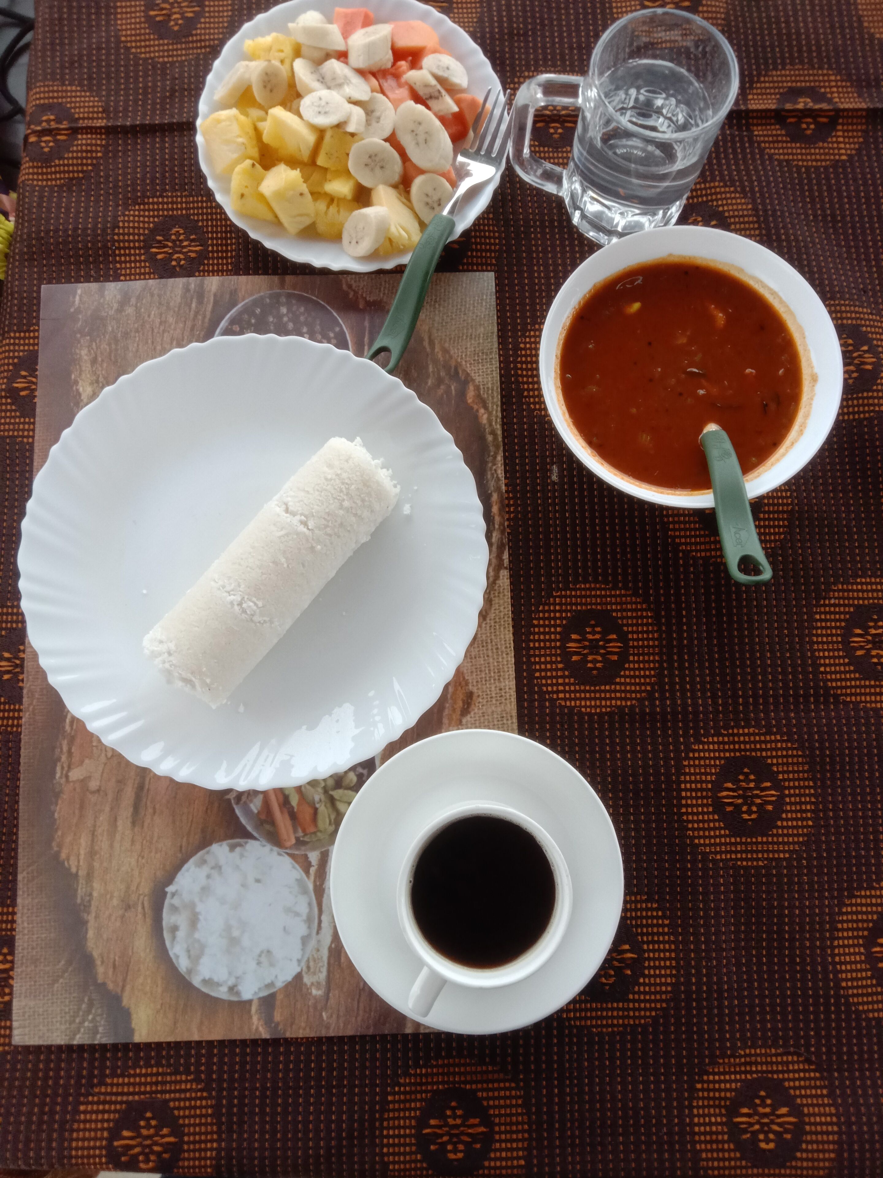 A meal with a plate of white food, a bowl of fruit, a bowl of soup, a cup of coffee, and a glass of water on a patterned tablecloth.