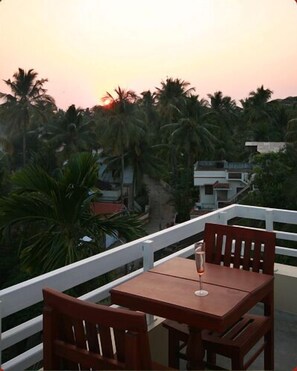A terrace with two wooden chairs and a table, overlooking a row of palm trees and buildings during sunset. - Orion Sky Wings (Kochi)