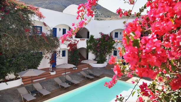 A poolside area with lounge chairs and umbrellas, surrounded by white buildings with blue doors and windows, and a mountainous backdrop. - Boutique Hotel Tilos Mare (Tilos)
