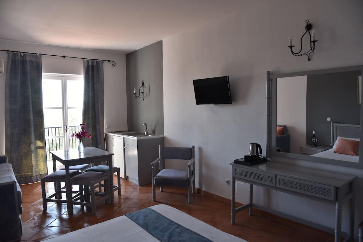 Une chambre à coucher comprenant un lit, un bureau, une chaise, une télévision, un miroir et un balcon avec des rideaux.