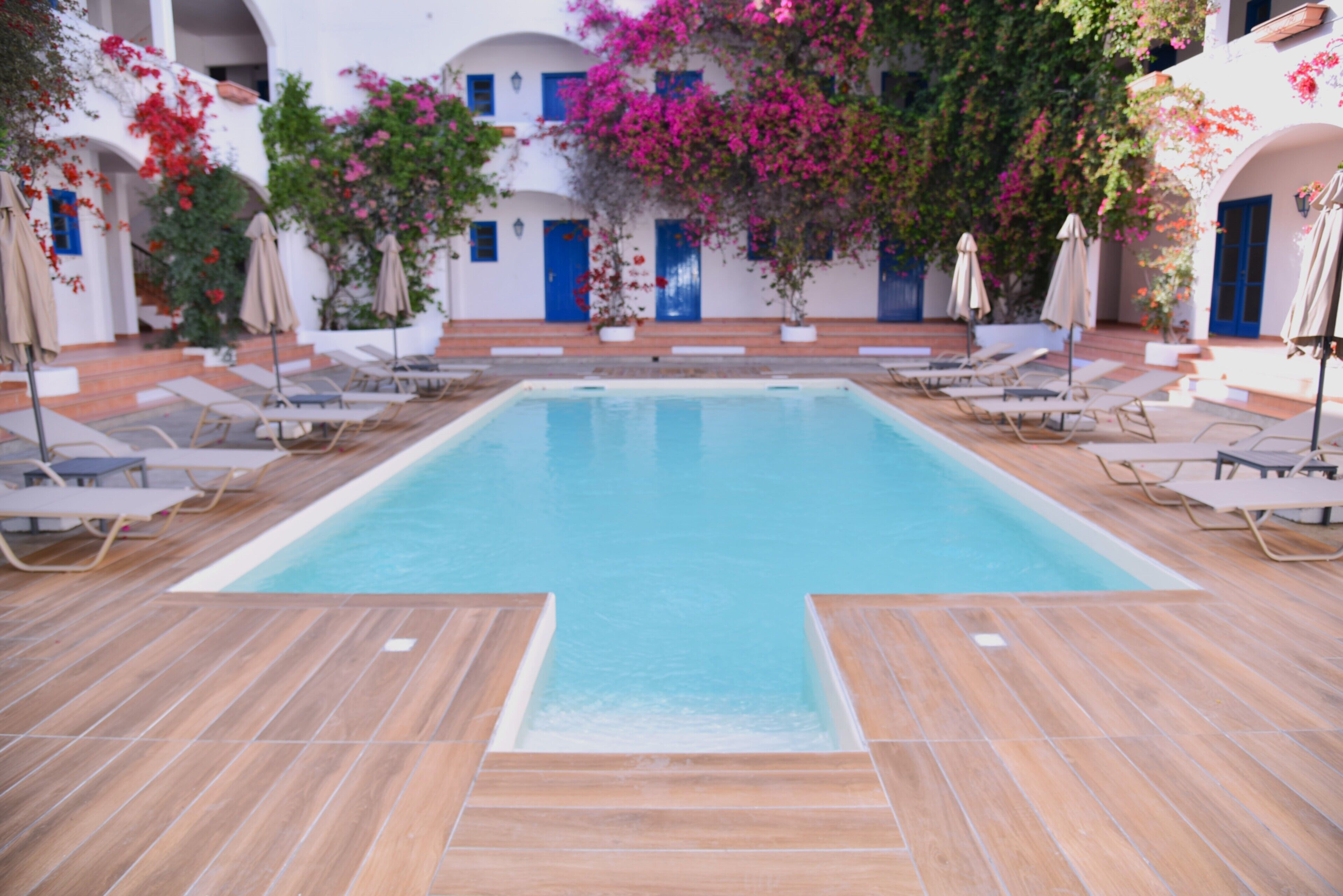 A swimming pool with lounge chairs and umbrellas, surrounded by white buildings with blue doors and pink flowering plants.