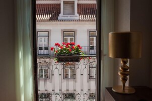 A window with a view of a building's facade, a balcony with red flowers, and a lamp on a table.