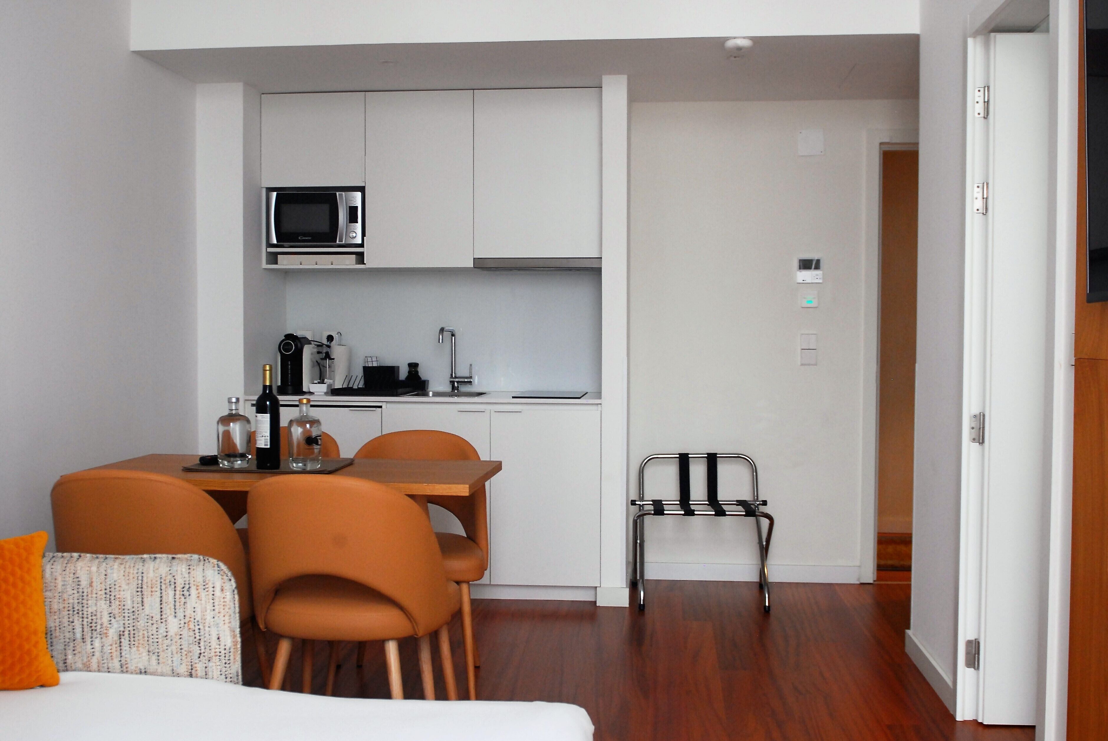 Une chambre d’hôtel compacte comprenant un coin cuisine, un espace repas avec une table et des chaises, et un lit agrémenté d’un oreiller orange.