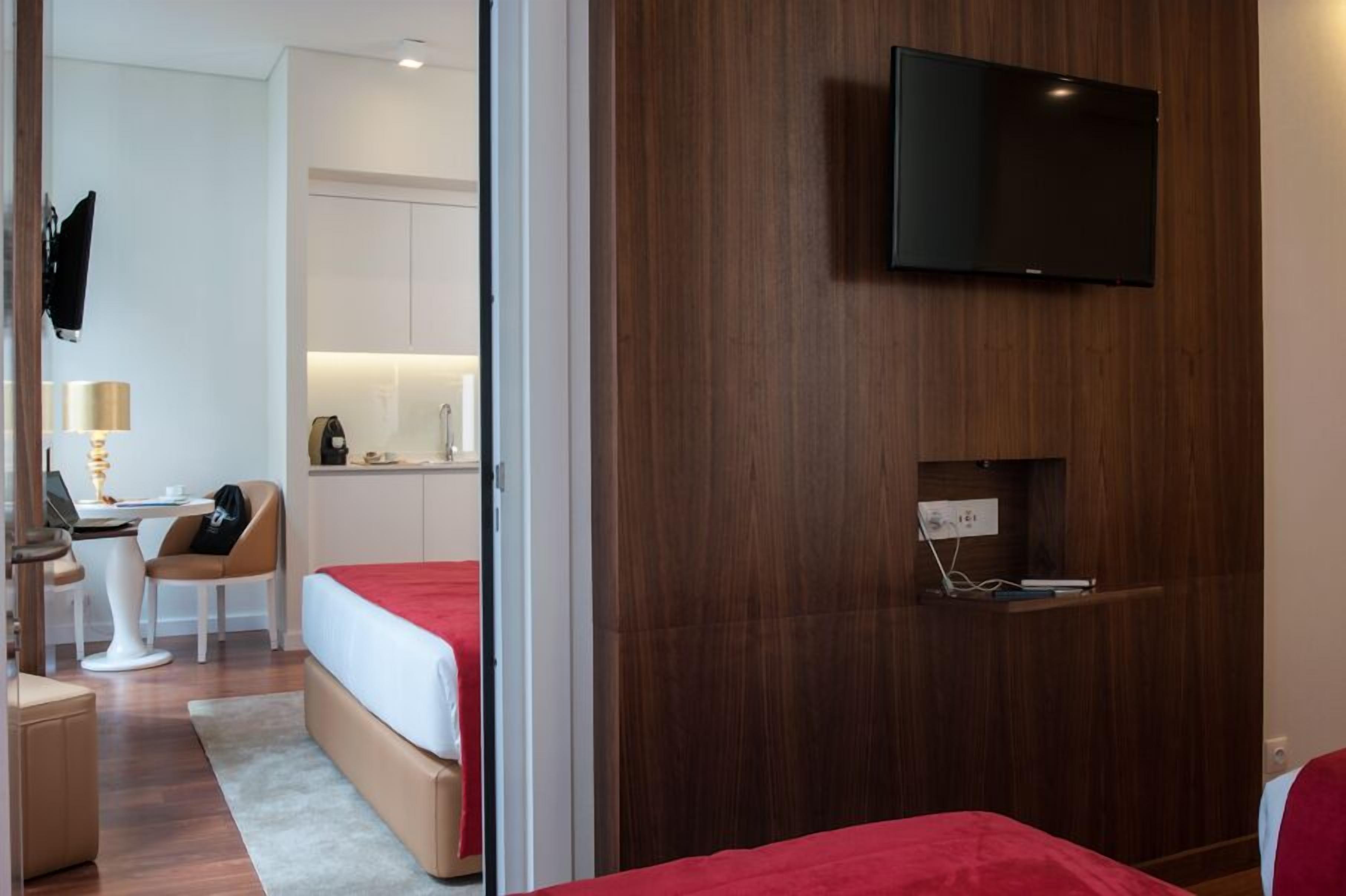 A hotel room with a bed, a desk with a chair, a television mounted on the wall, and a small shelf with items.