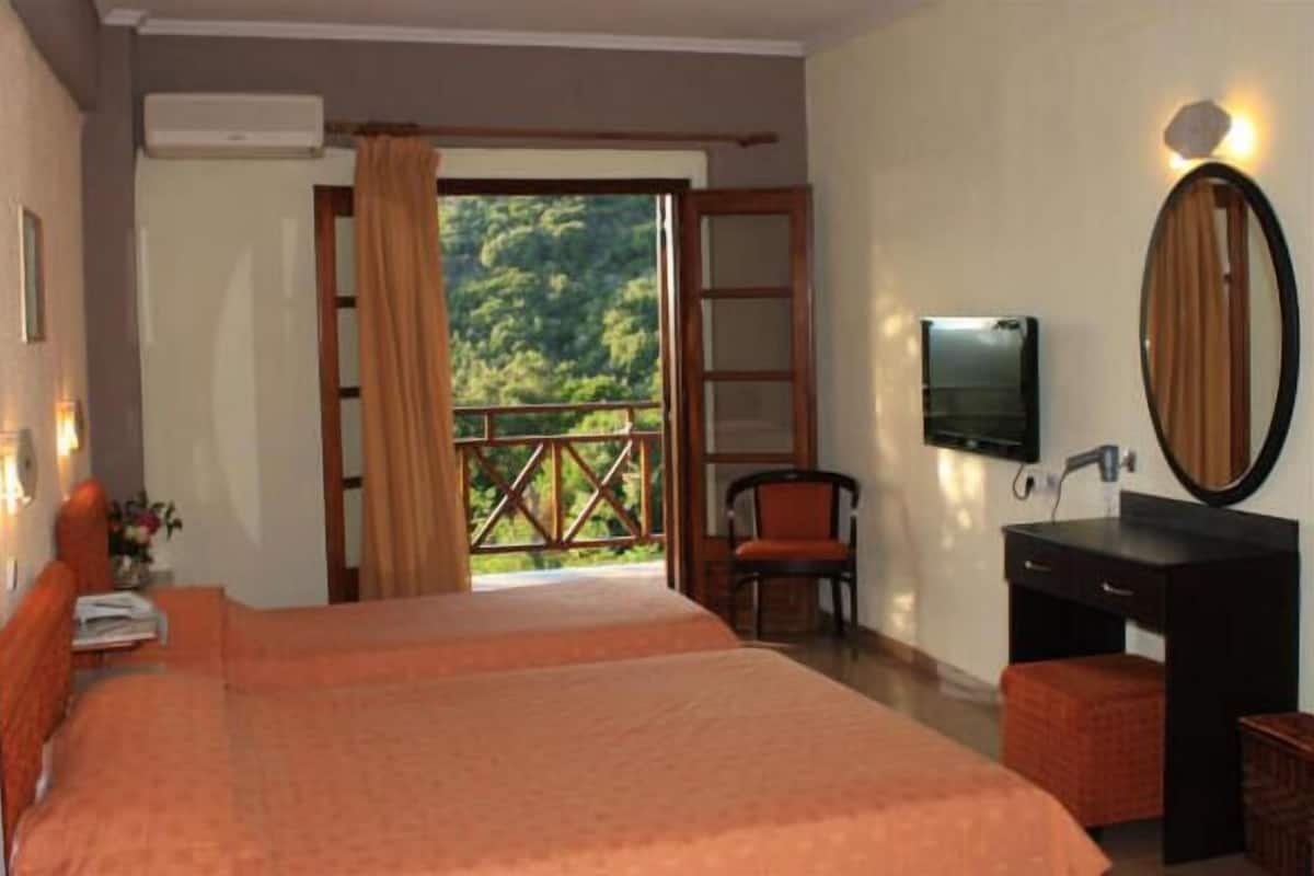 A hotel room with a bed, a desk with a mirror, a chair, and a TV. There is a balcony with a view of greenery.