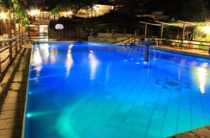 Una piscina con aro de baloncesto, rodeada por una terraza de madera y un muro de piedra.