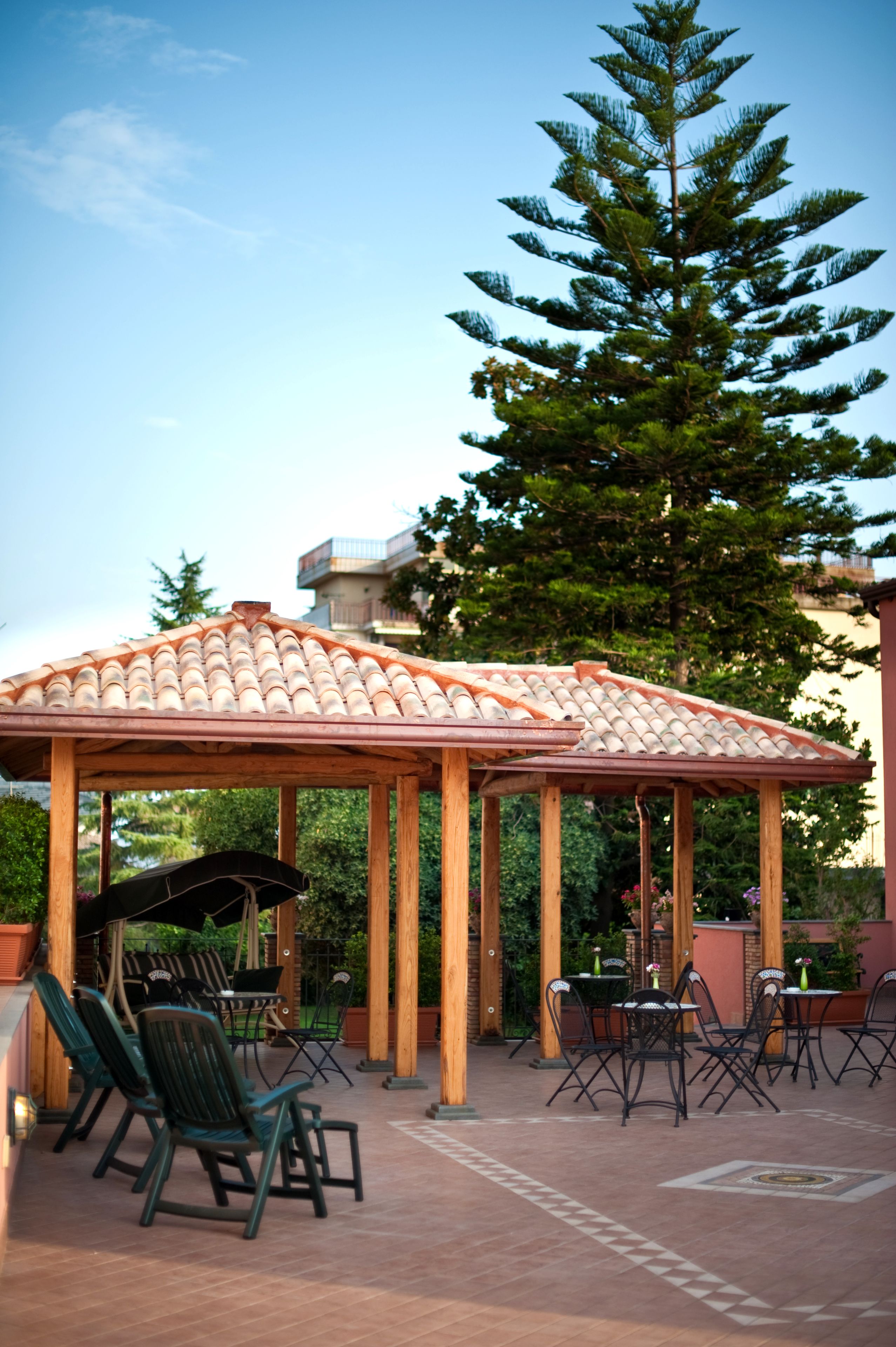 Ein überdachter Patio-Bereich mit Holztischen und -stühlen, umgeben von viel Grün und einem hohen Baum.