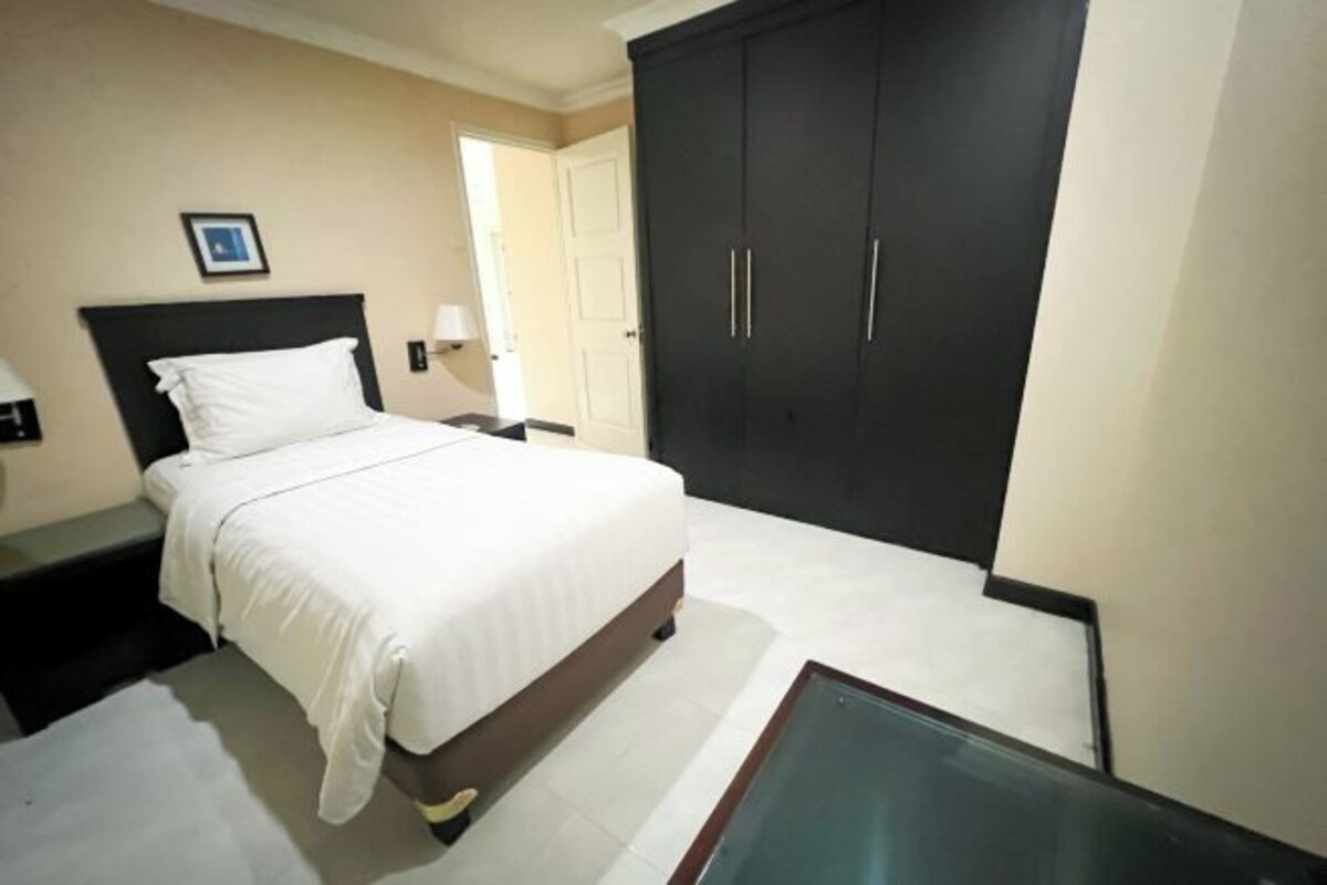 A hotel room with a bed, a nightstand, a desk, a wardrobe, and a glass table.