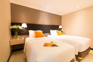 Habitación de hotel con dos camas, cada una con ropa de cama blanca y almohadas naranjas. Se ve una mesita de noche con una lámpara y un jarrón de flores a la izquierda.
