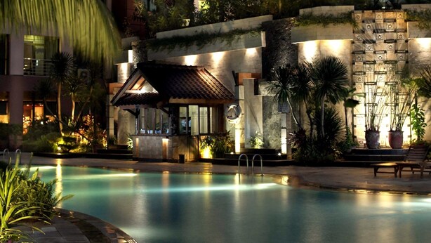 Un área de piscina con un edificio de estilo tradicional, un muro de piedra con diseños decorativos y abundante vegetación.