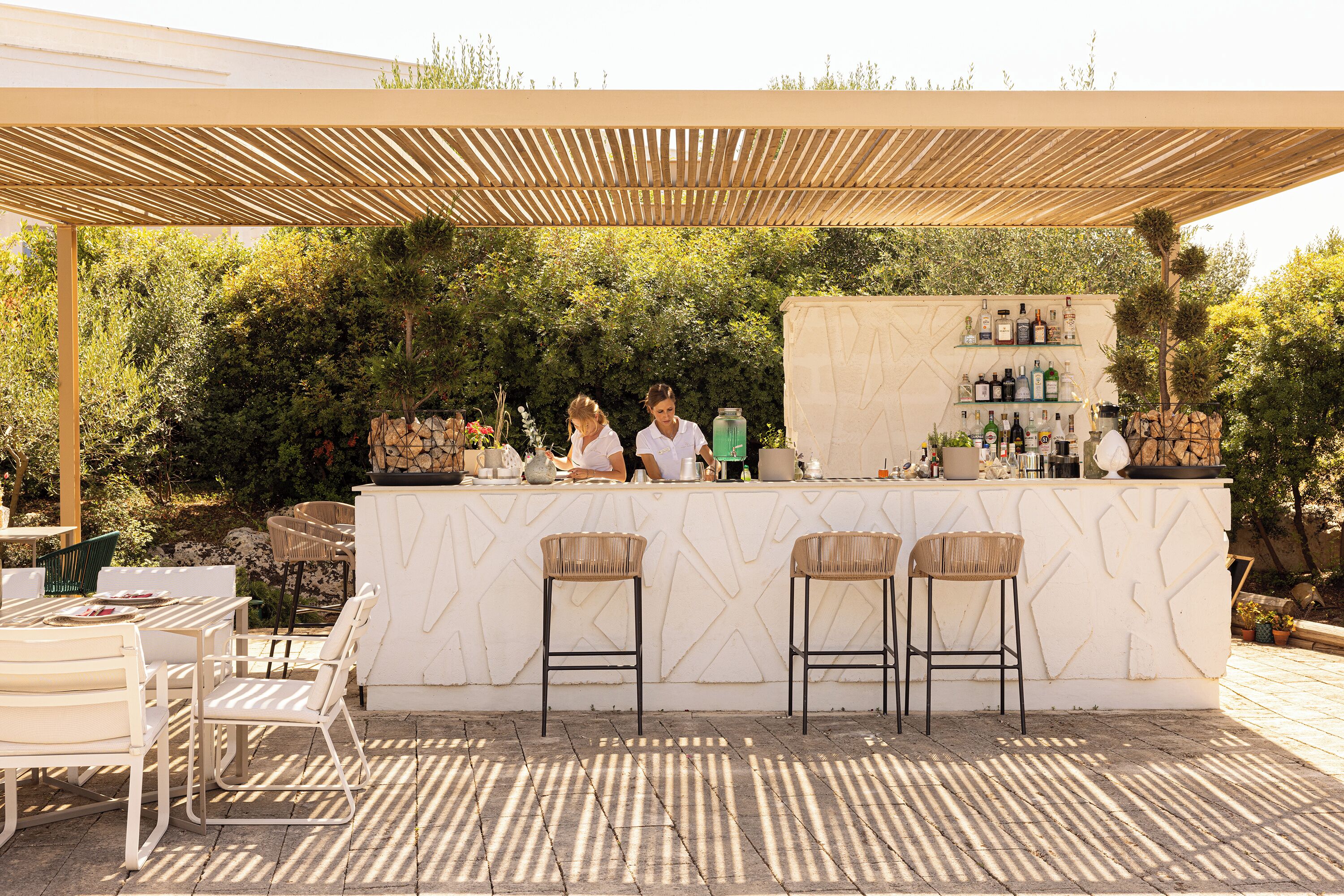 Un espace bar extérieur doté d’un comptoir en marbre blanc, de tabourets de bar en osier et d’une pergola en bois au-dessus.