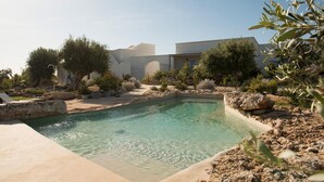 A pool area with clear water, surrounded by rocks and plants, and a building in the background. - Masseria San Francesco (Fasano)