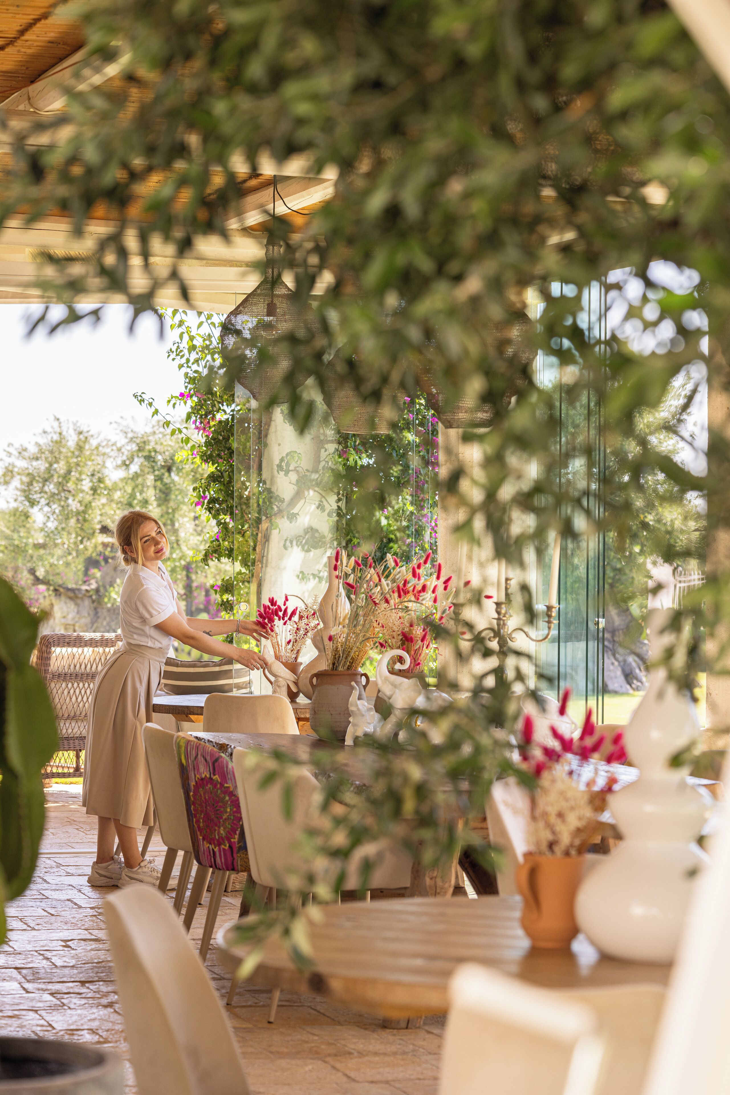 A woman arranging flowers on a table in an outdoor setting with a glass partition and greenery.