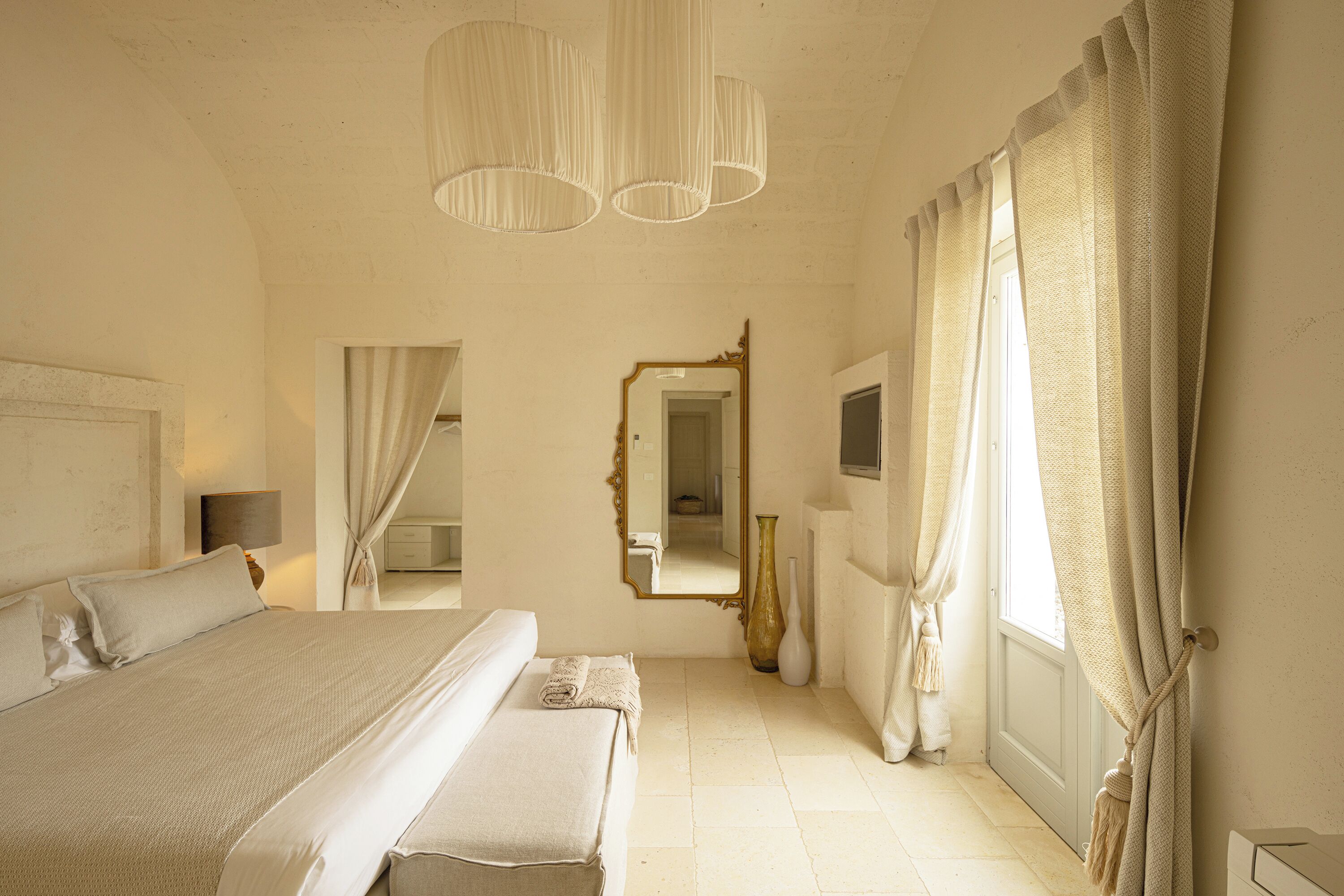A bedroom with a large bed, a mirror, a vase, and curtains.