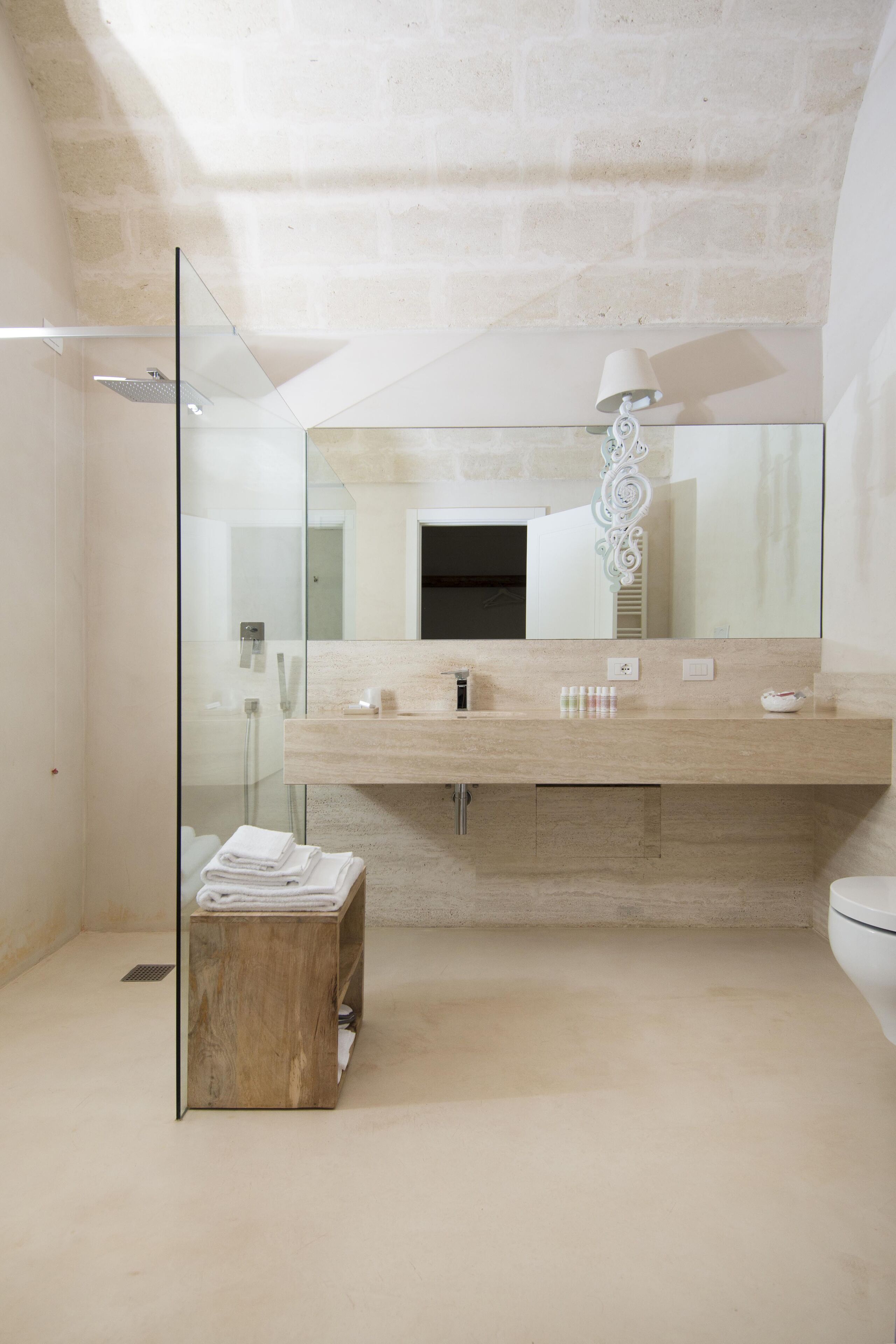 Et moderne badeværelse med brusekabine i glas, træbænk, stort spejl og bordplade i beige med toiletartikler.