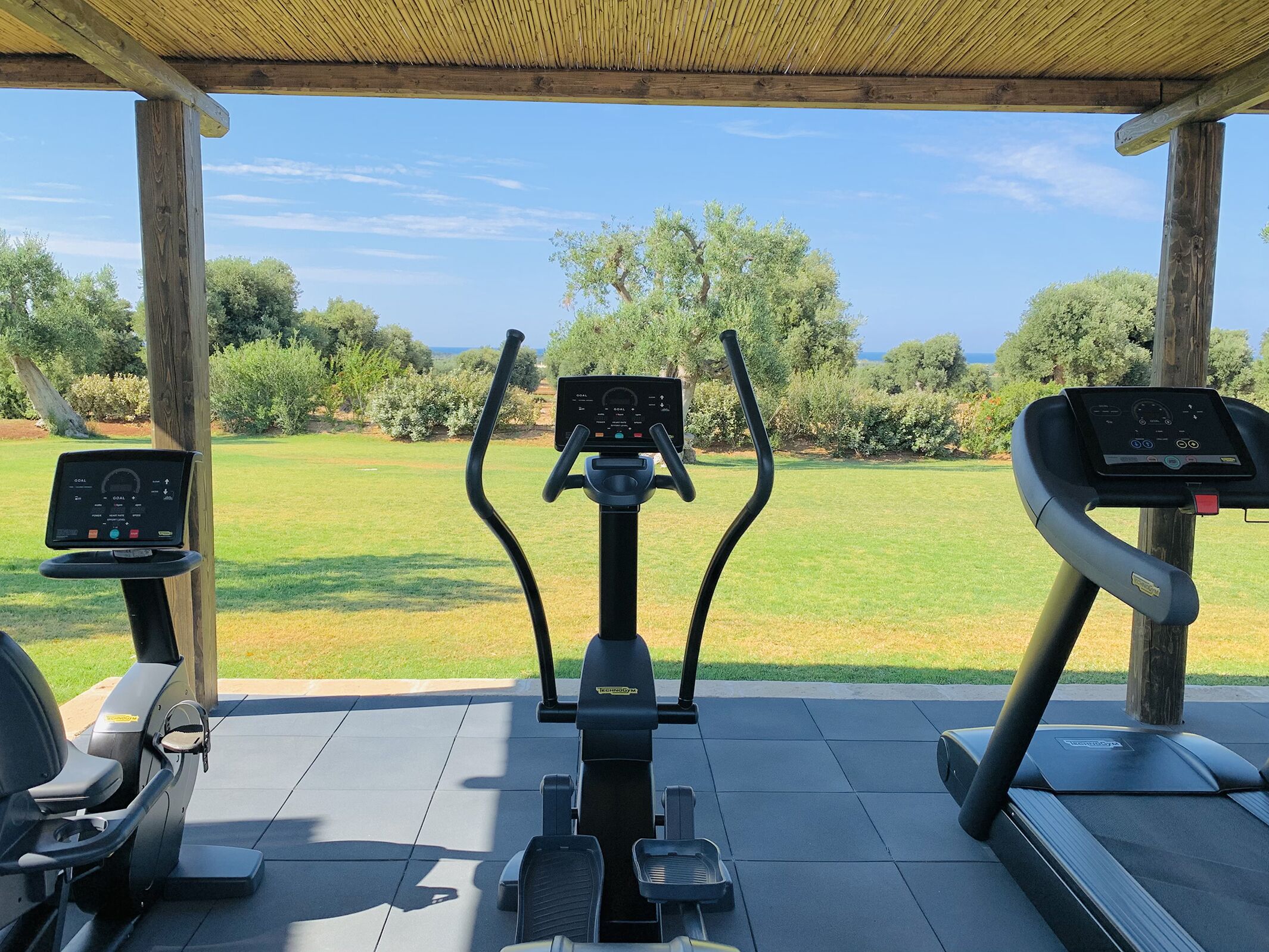An outdoor gym area with exercise equipment including treadmills and an elliptical machine, under a wooden pergola with a view of a grassy field and trees.
