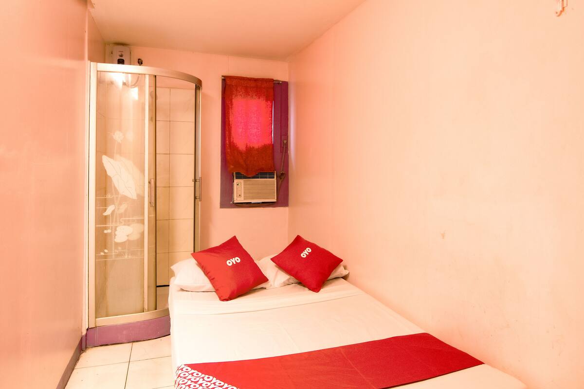 A small, compact bedroom with a bed, two red pillows, a red blanket, a shower, and a window with a red curtain.