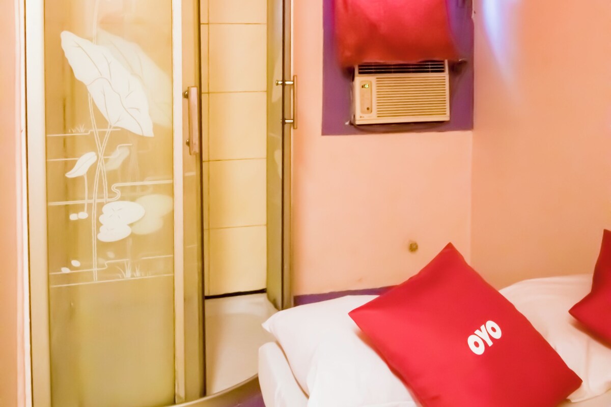 A bedroom with a bed, two red pillows, a glass shower enclosure, and a wall-mounted air conditioning unit.
