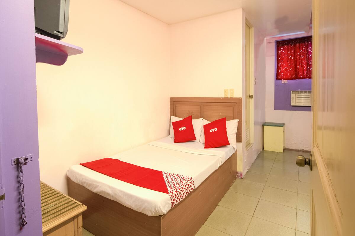 A small, neatly arranged bedroom with a bed, a bedside table, a television on a shelf, and a window with red curtains.