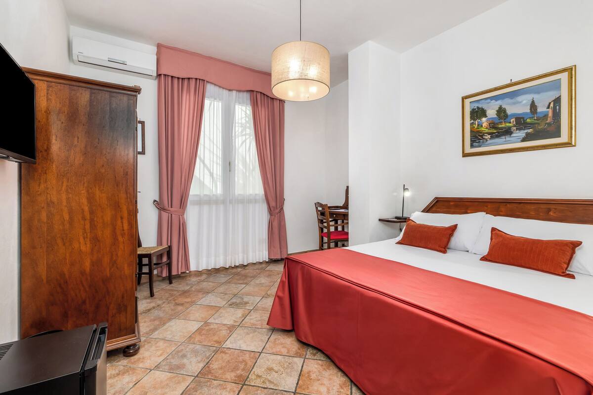 A hotel room with a bed, a wooden wardrobe, a television, a painting on the wall, and a window with curtains.