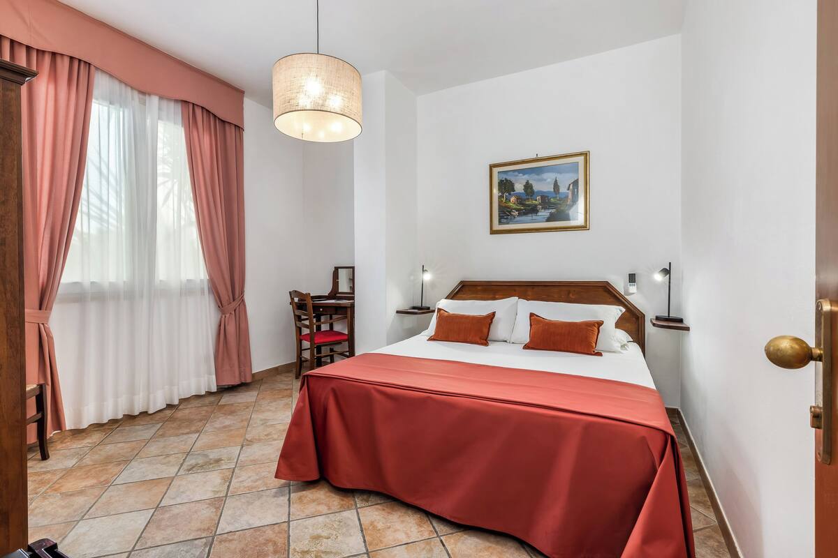 A hotel room with a bed, a chair, a desk, a painting on the wall, and a window with curtains.