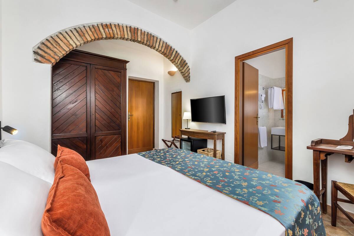 A hotel room with a bed, a TV, a desk, a wardrobe, and an arched doorway.