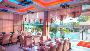 A restaurant with tables set for dining, large windows with blinds, and a view of the sea.