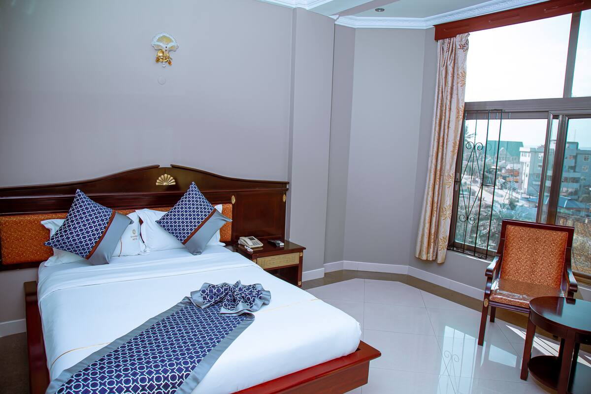 A hotel room with a large bed, a nightstand, a chair, and a view of the city through a large window.