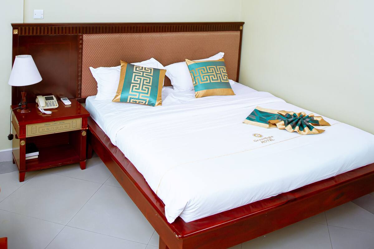 A hotel room with a wooden bed, white bedding, a nightstand with a lamp, and a wall-mounted air conditioner.