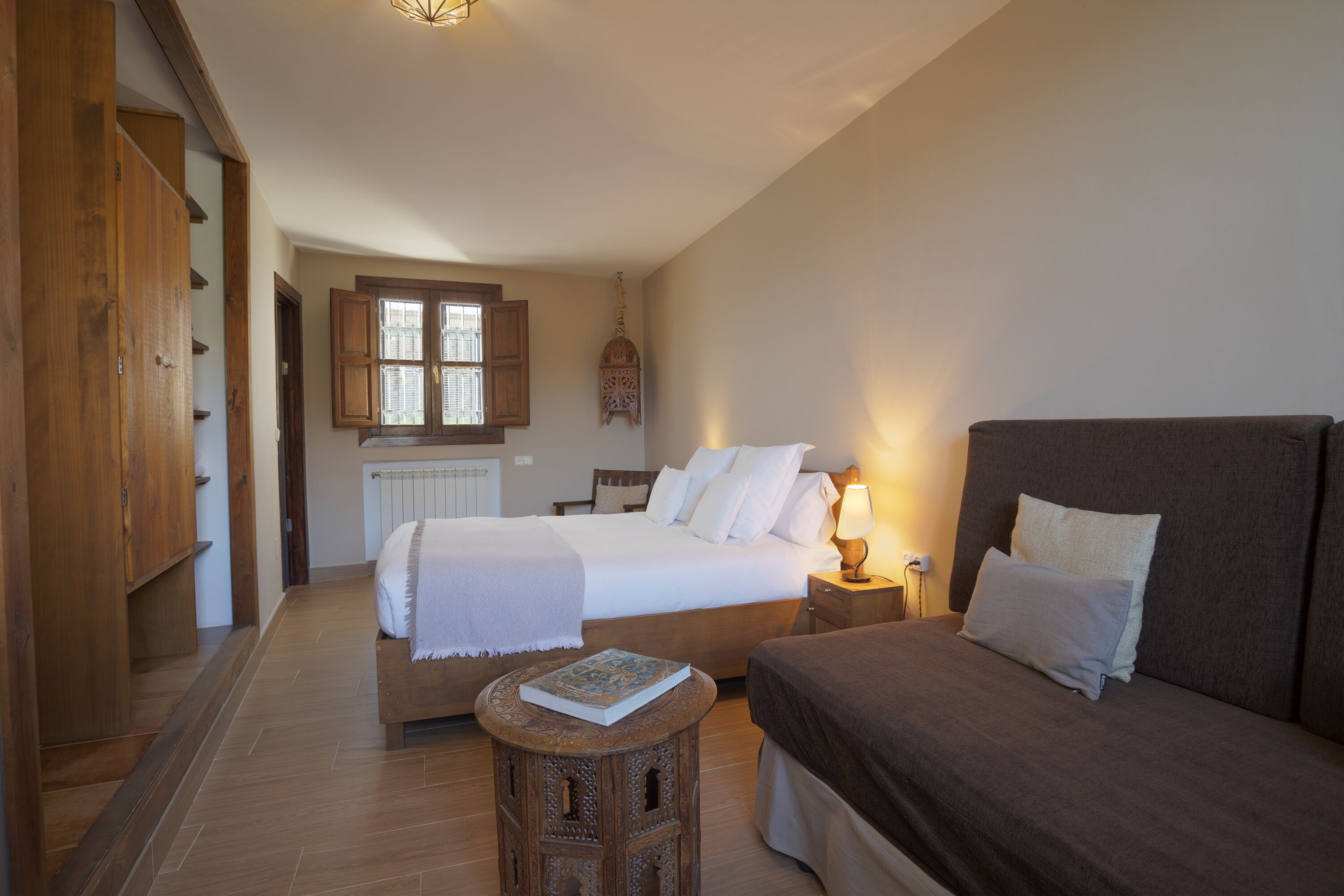 A bedroom with a large bed, a smaller bed, a wooden wardrobe, a nightstand with a lamp, and a decorative hanging light.