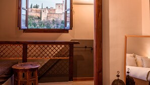 A bedroom with a bed, a window with a view of a cityscape, a wooden floor, and a small round table. - Casa Bombo (Granada)