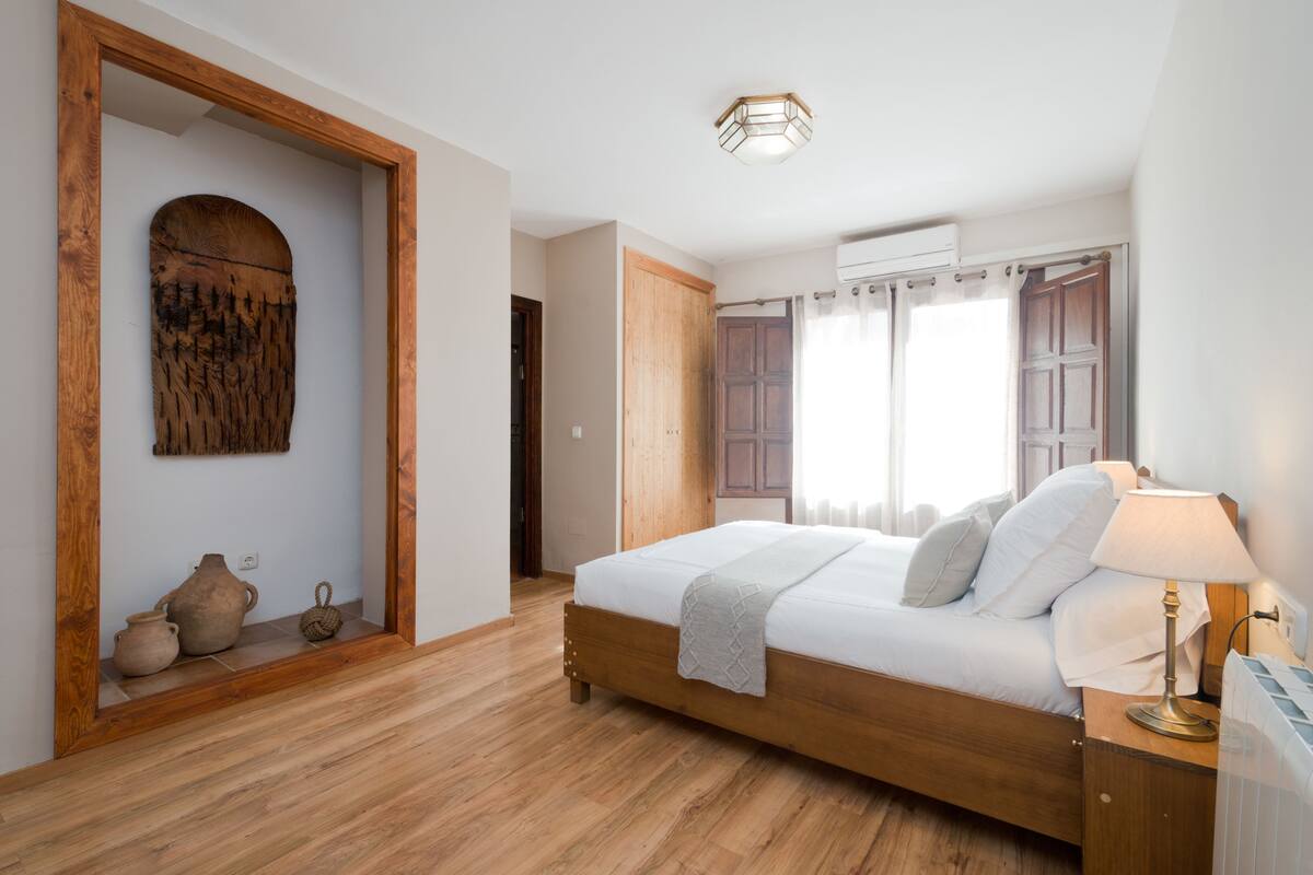 A bedroom with a wooden bed, a nightstand, a lamp, a window with curtains, and a wooden door.