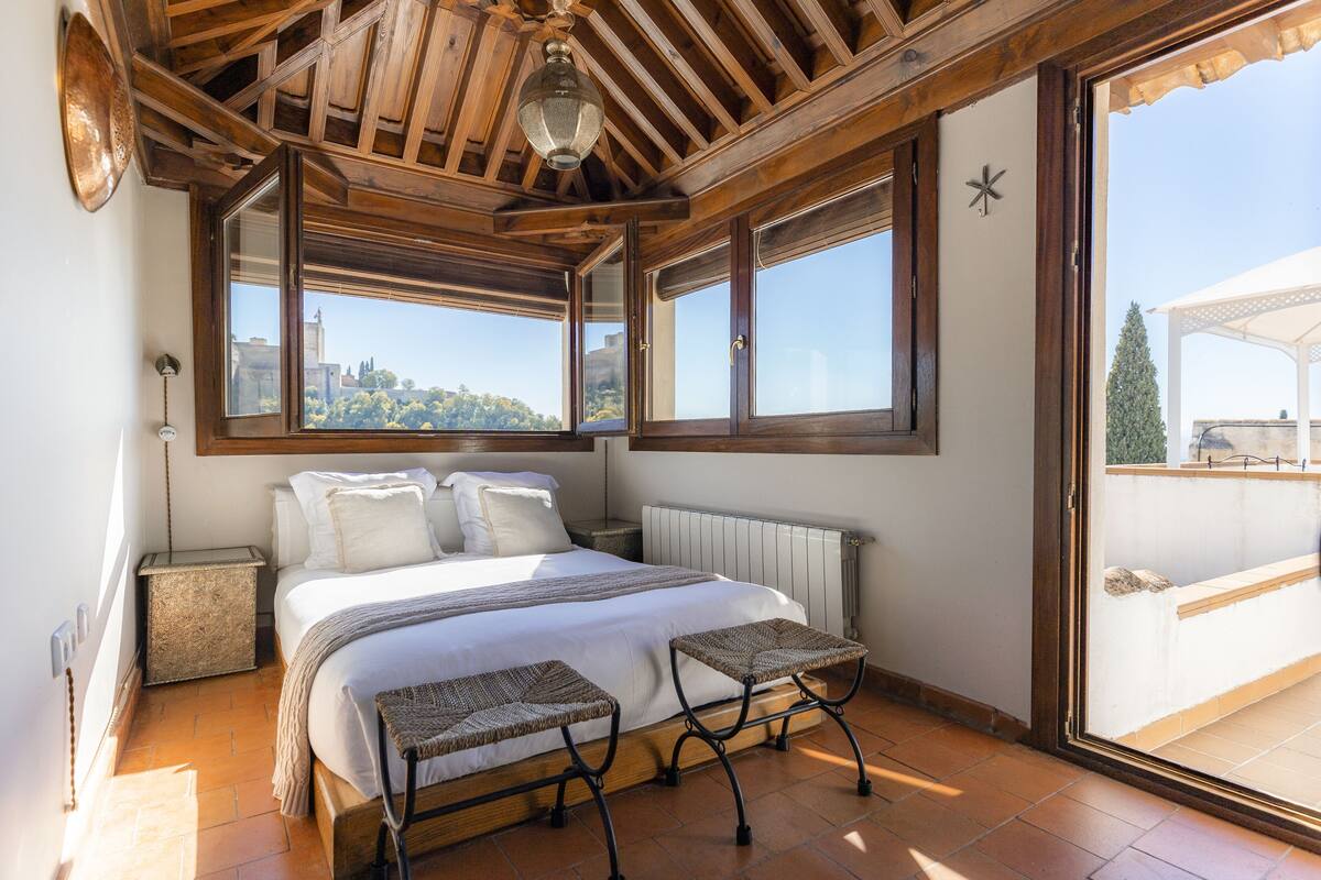 A bedroom with a large bed, two woven stools, a radiator, and a city view through large windows.