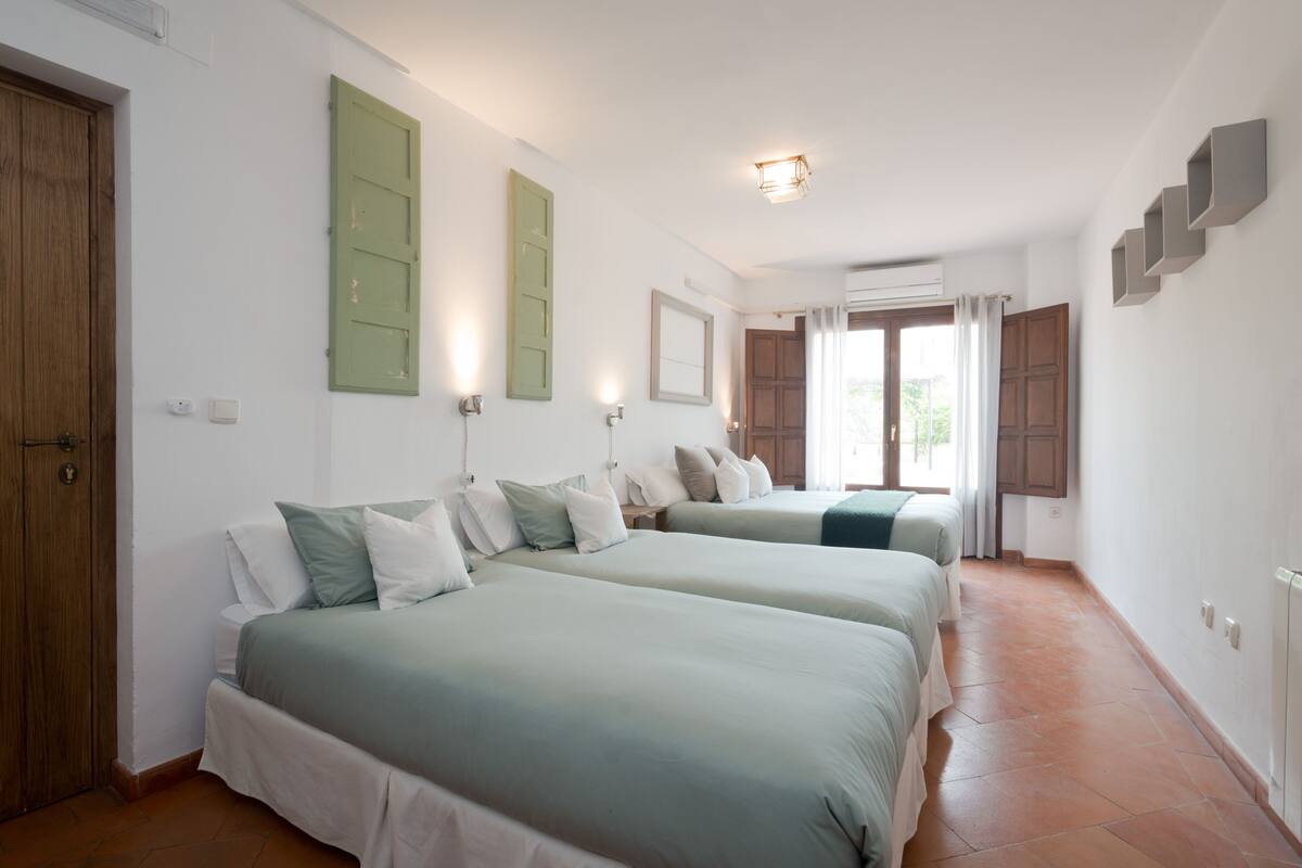 A hotel room with two beds, a wooden door, a window with curtains, and a tiled floor.
