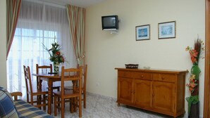 Una sala de estar con sofá, mesa de comedor, sillas, televisor, aparador de madera y un florero con flores.