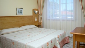 Una habitación de hotel con cama, cabecero de madera, mesita de noche, lámpara y ventana con cortinas.