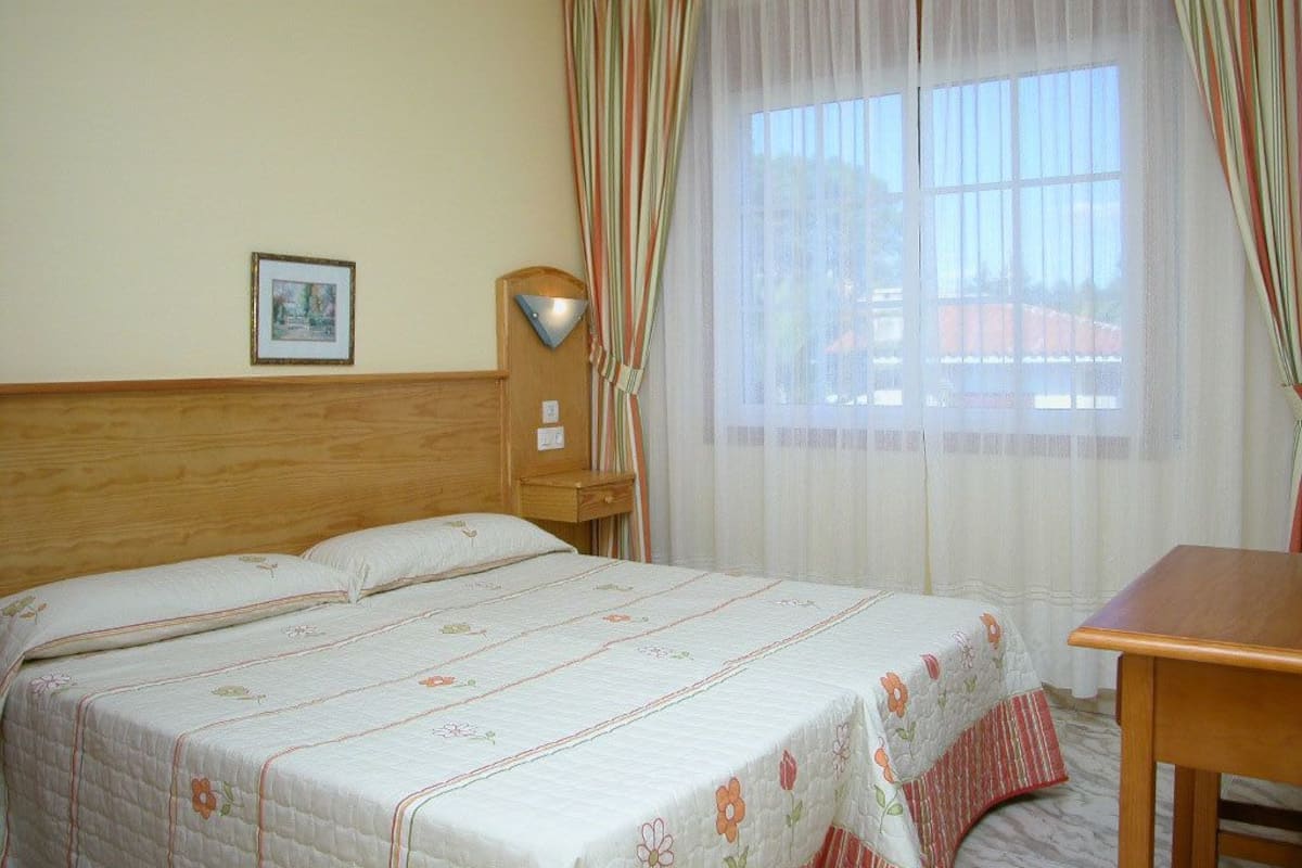 Une chambre d’hôtel avec un lit, une tête de lit en bois, une table de chevet, une lampe et une fenêtre avec des rideaux.