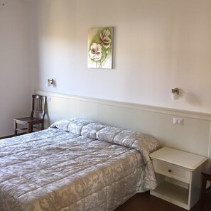 A bedroom with a bed, a chair, a nightstand, and a wall-mounted light switch.