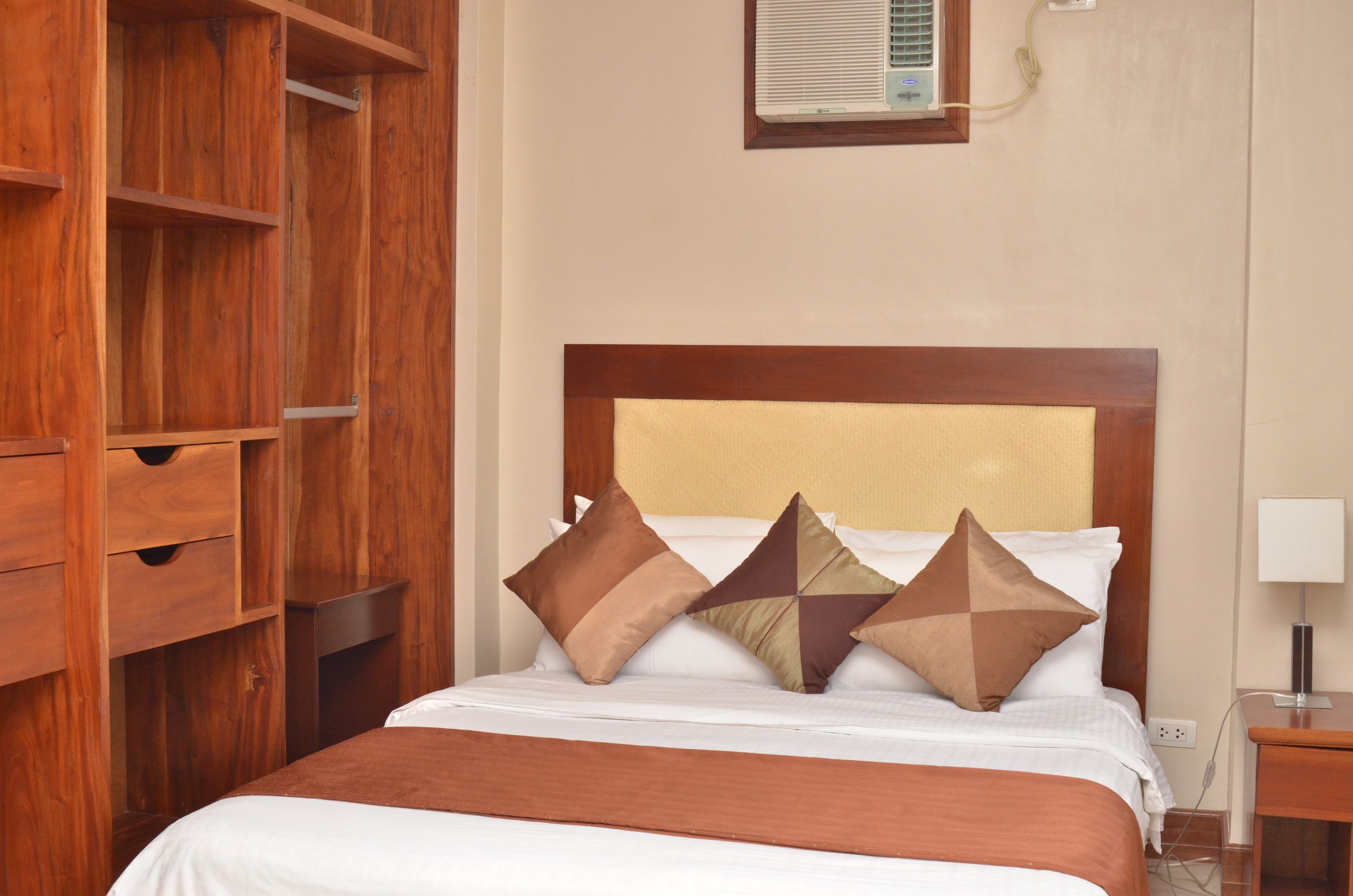 A hotel room with a wooden wardrobe, a bed with white bedding and brown pillows, and a bedside table with a lamp.