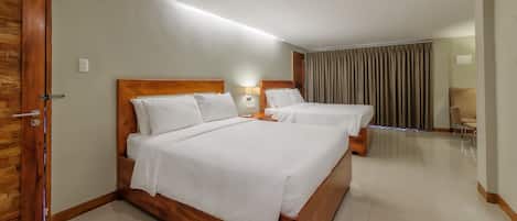 Habitación de hotel con dos camas, una puerta de madera, una mesita de noche, una lámpara y vistas al exterior a través de cortinas.