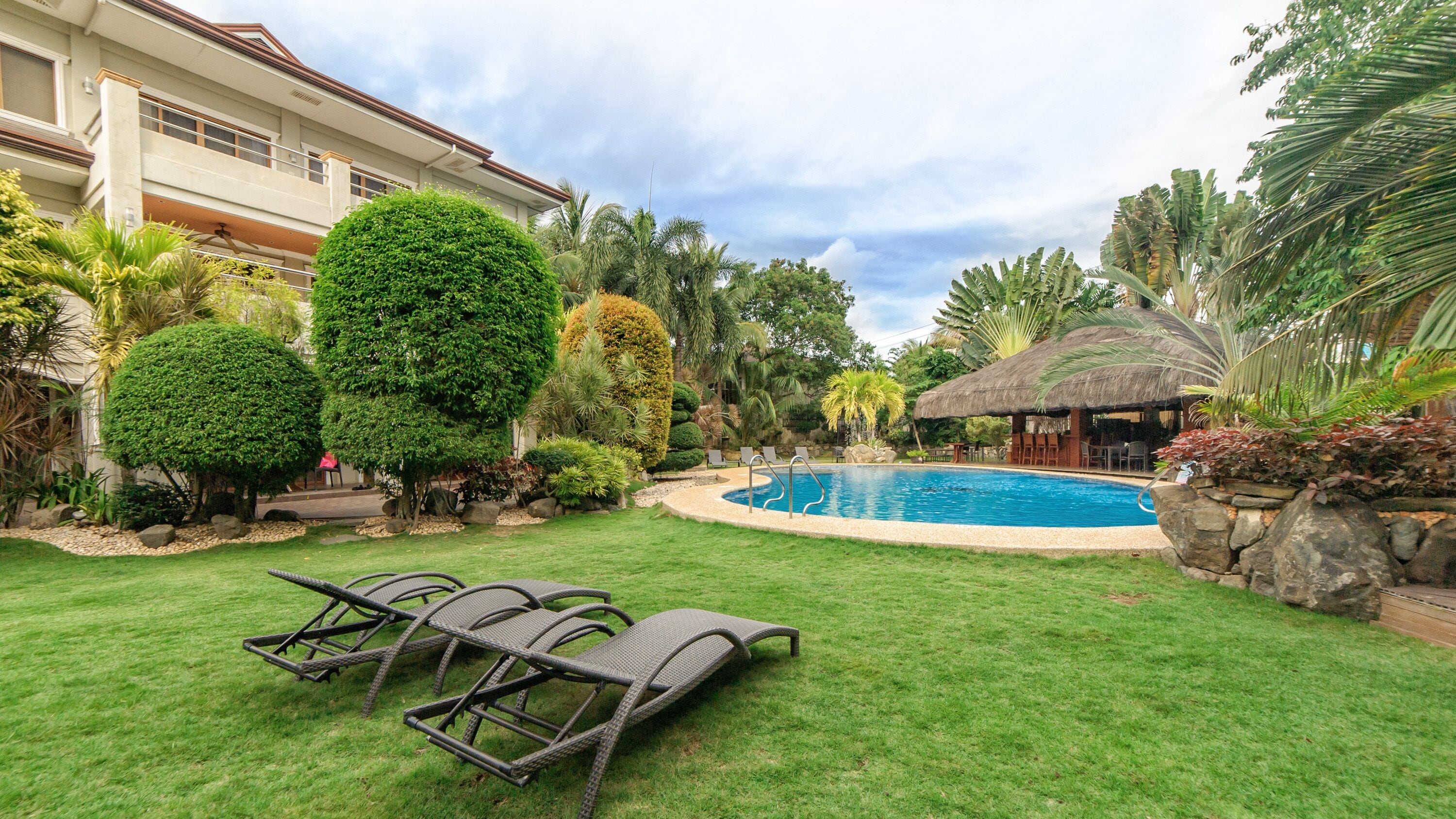 A swimming pool surrounded by lush greenery, a thatched-roof structure, and lounge chairs.
