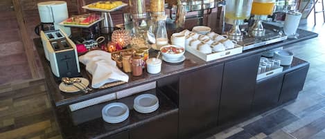 Uma mesa de buffet com diversos alimentos, utensílios de servir e distribuidores de bebidas.